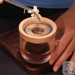 Pince déposant des copeaux de bois sur le filtre du fumoir en bois posé sur un verre Délice Bar