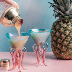 Deux verres méduse irisés bleus et roses, cocktail versé au shaker cuivré devant un ananas