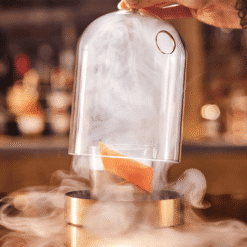 Grande cloche en verre à socle doré remplie de fumée et zeste d'orange en bar
