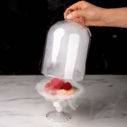 Main soulevant une cloche fumée révélant un dessert aux framboises sur présentoir en verre