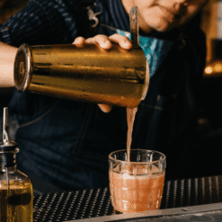 Barman versant un cocktail rosé d'un shaker Boston doré dans un verre tumbler