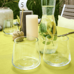 Deux gobelets Ava en verre transparent évasé sur nappe jaune avec carafe d'eau aromatisée, table dressée