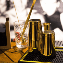 Shaker Boston doré Kenko et doseur dorés sur tapis de bar, cuillère et verre à mélange en arrière-plan