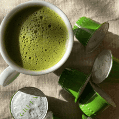 Tasse de matcha latte vert mousseux entouree de capsules vertes Green ouvertes sur toile beige