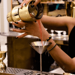 Versement d'un cocktail rose depuis le shaker Parisien Zeus dore vers un verre martini sur le bar