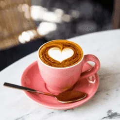 Tazza rosa di cappuccino con cuore disegnato in latte art su tavolo di marmo e cucchiaino