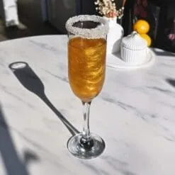 Flûte con cocktail dorato glitterato dal bordo ghiacciato di zucchero su un tavolo di marmo illuminato dal sole