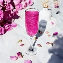 Flûte con cocktail rosa fucsia scintillante circondata da rose e boccioli essiccati, glitter alimentare glamour