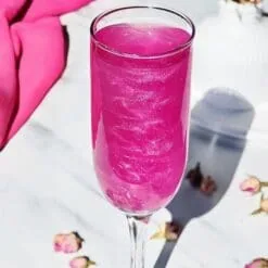Flûte de cocktail rose fushia scintillant sur marbre avec boutons de roses séchés, paillettes comestibles