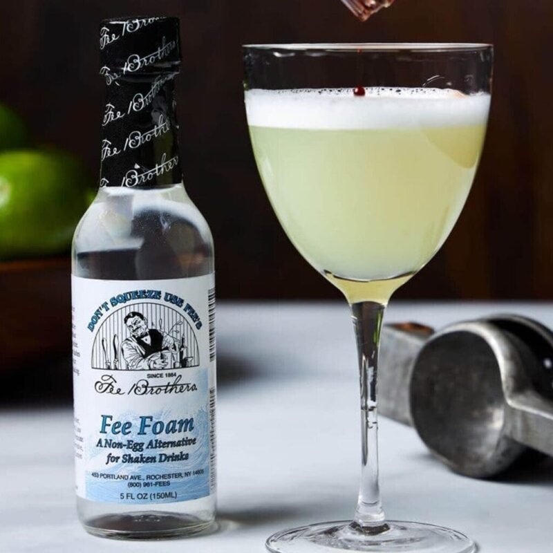 Cocktail sour chiaro in calice, con schiuma bianca punteggiata da una goccia di bitter, accanto a un flacone di Fee Foam
